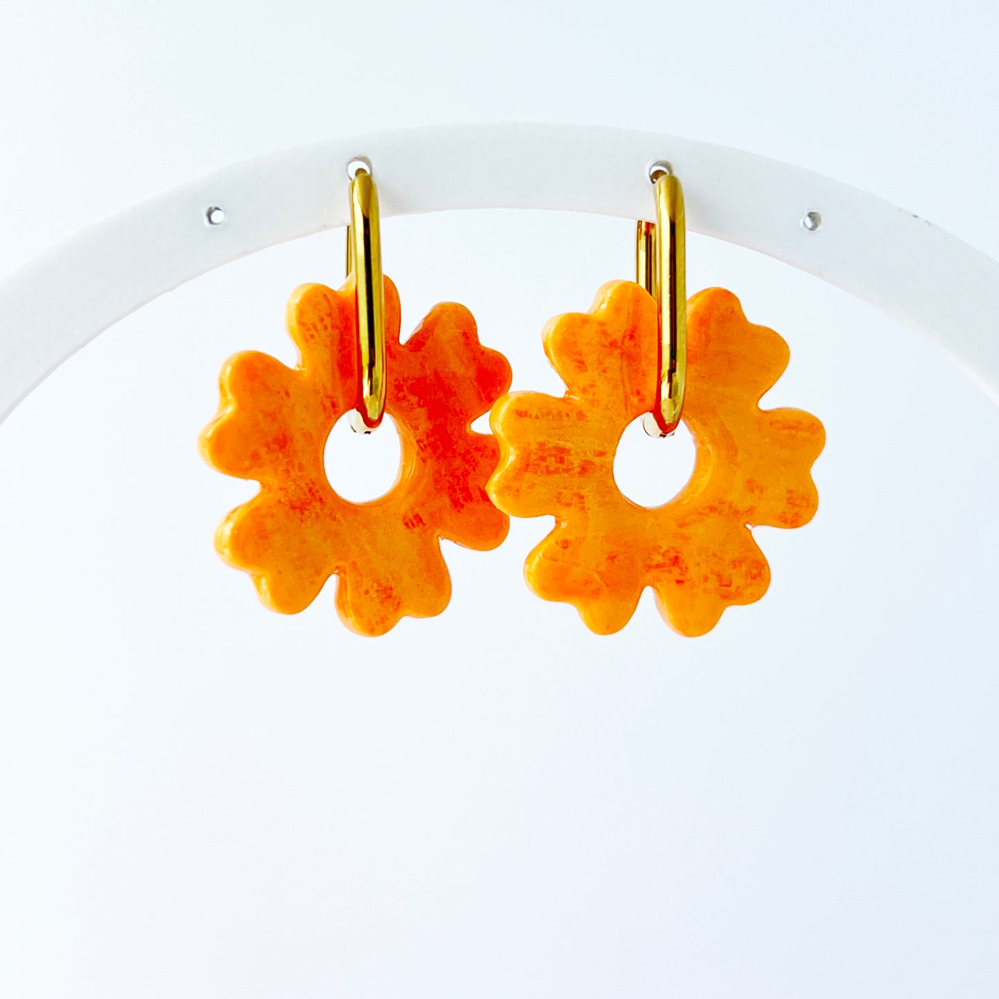 A pair of orange handmade polymer clay earrings shaped like abstract flowers, each dangling from a gold oval-shaped 316L stainless steel hoop. 