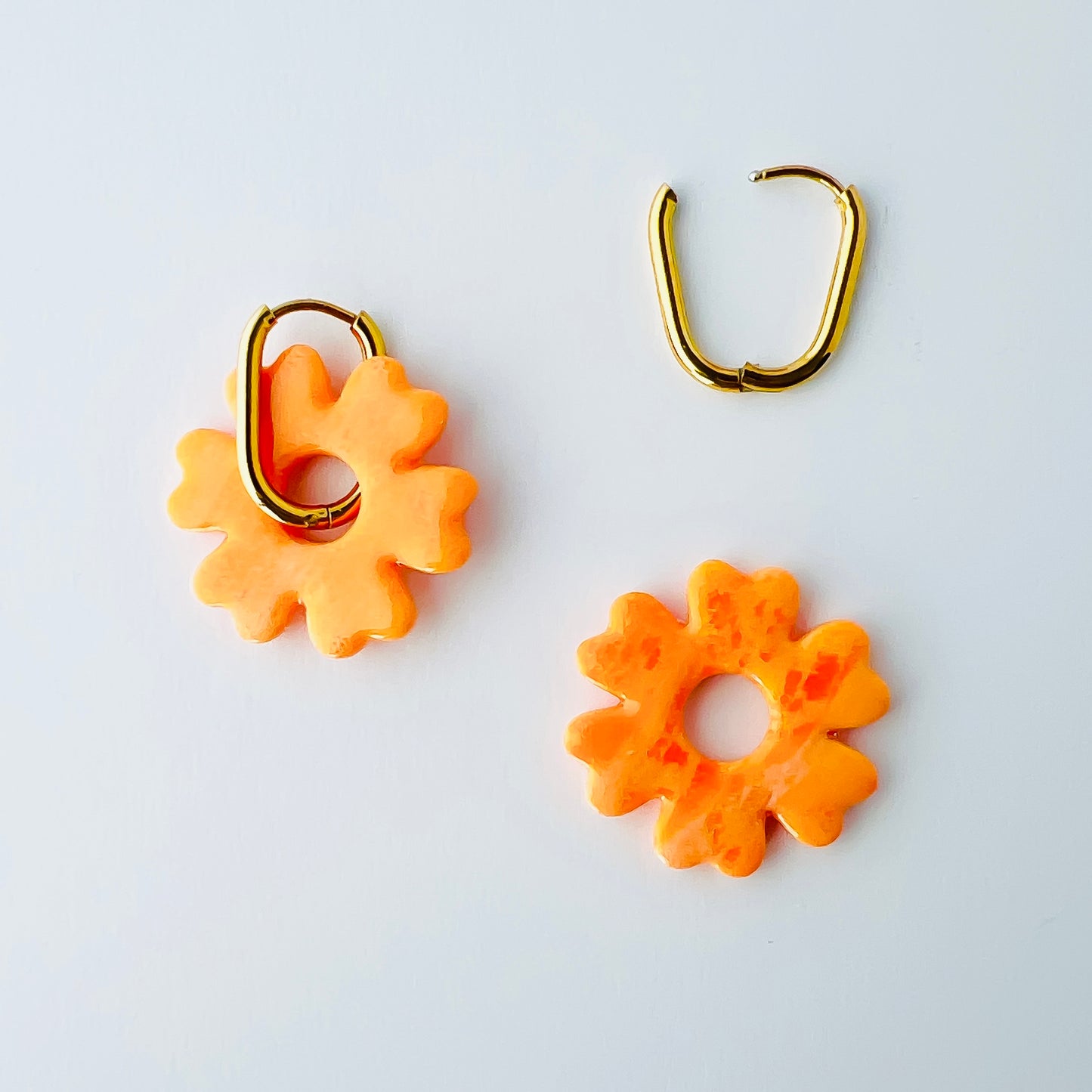  A pair of orange handmade polymer clay earrings shaped like abstract flowers, each dangling from a gold oval-shaped 316L stainless steel hoop. One half of the photo shows the hoops worn with the flower, and another shows the flower removed to demonstrate the 2-in-1 styling option.
