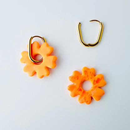  A pair of orange handmade polymer clay earrings shaped like abstract flowers, each dangling from a gold oval-shaped 316L stainless steel hoop. One half of the photo shows the hoops worn with the flower, and another shows the flower removed to demonstrate the 2-in-1 styling option.