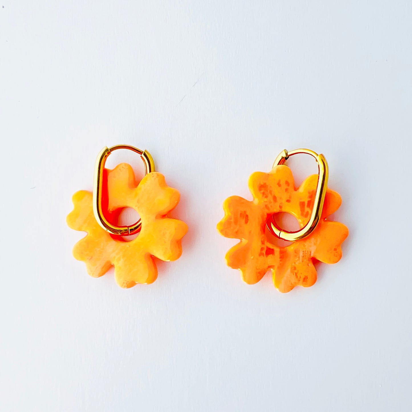 A pair of orange handmade polymer clay earrings shaped like abstract flowers, each dangling from a gold oval-shaped 316L stainless steel hoop. 