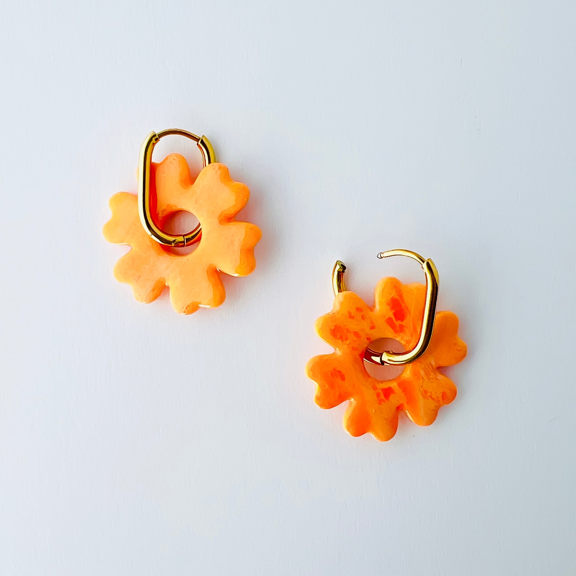  A pair of orange handmade polymer clay earrings shaped like abstract flowers, each dangling from a gold oval-shaped 316L stainless steel hoop. 