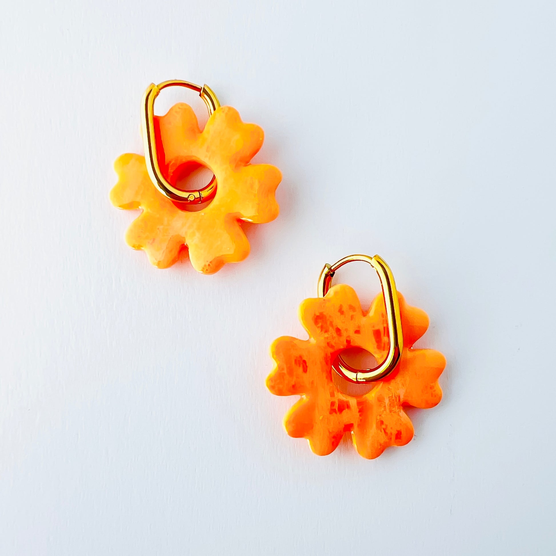  A pair of orange handmade polymer clay earrings shaped like abstract flowers, each dangling from a gold oval-shaped 316L stainless steel hoop. 