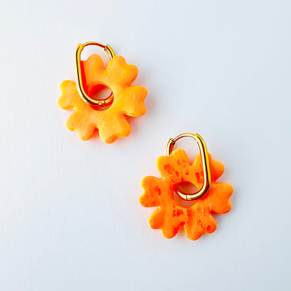  A pair of orange handmade polymer clay earrings shaped like abstract flowers, each dangling from a gold oval-shaped 316L stainless steel hoop. 