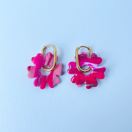 A pair of bold pink handmade polymer clay earrings shaped like abstract flowers, each dangling from a gold oval-shaped 316L stainless steel hoop. 