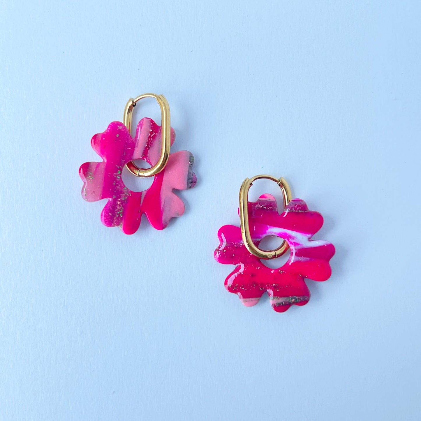 A pair of bold pink handmade polymer clay earrings shaped like abstract flowers, each dangling from a gold oval-shaped 316L stainless steel hoop. 