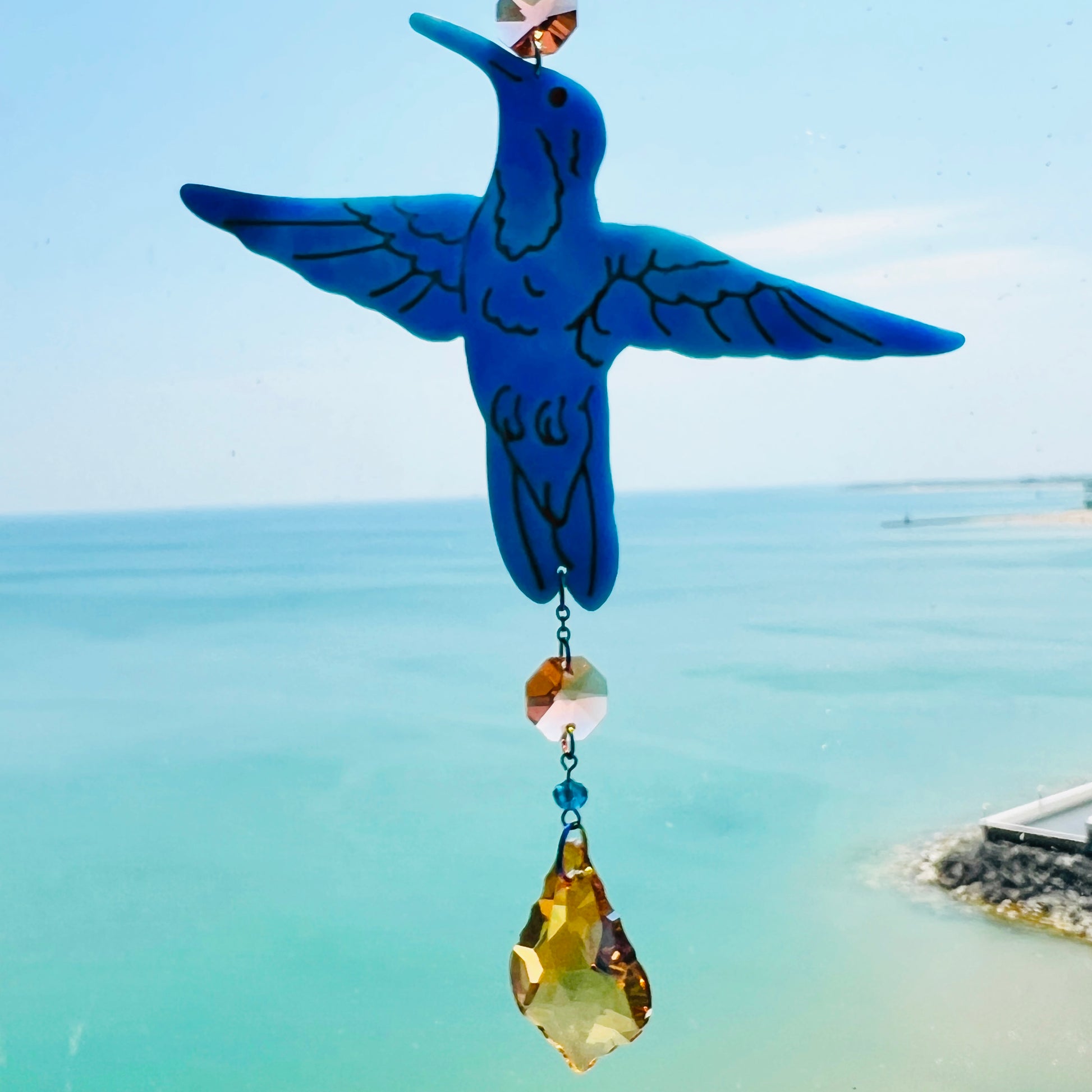 A handmade hummingbird suncatcher crafted from semi-transparent blue polymer clay, featuring a detailed hummingbird design suspended from a gold chain with a faceted champagne maple leaf glass crystal that casts rainbows in the sunlight. Perfect for nature lovers and whimsical window decor.