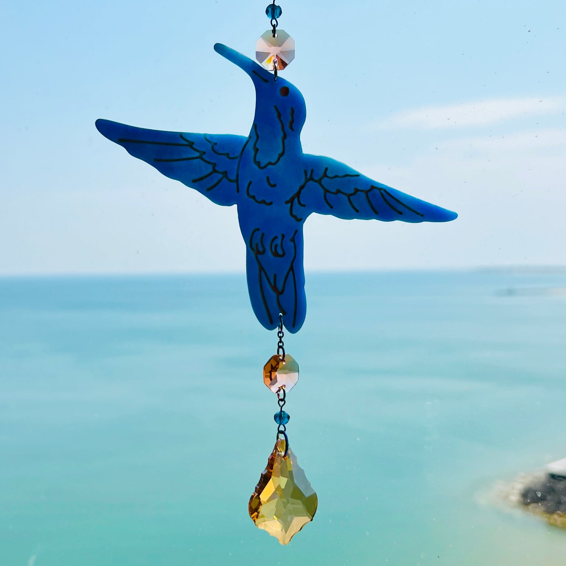 A handmade hummingbird suncatcher crafted from semi-transparent blue polymer clay, featuring a detailed hummingbird design suspended from a gold chain with a faceted champagne maple leaf glass crystal that casts rainbows in the sunlight. Perfect for nature lovers and whimsical window decor.