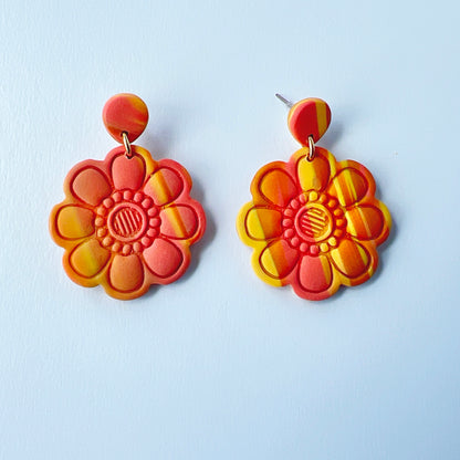 Handmade polymer clay earrings shaped like large, layered flowers with nine sculpted petals in a marbled blend of coral, orange, and yellow tones, finished with textured flower centers and coral clay stud tops on titanium posts