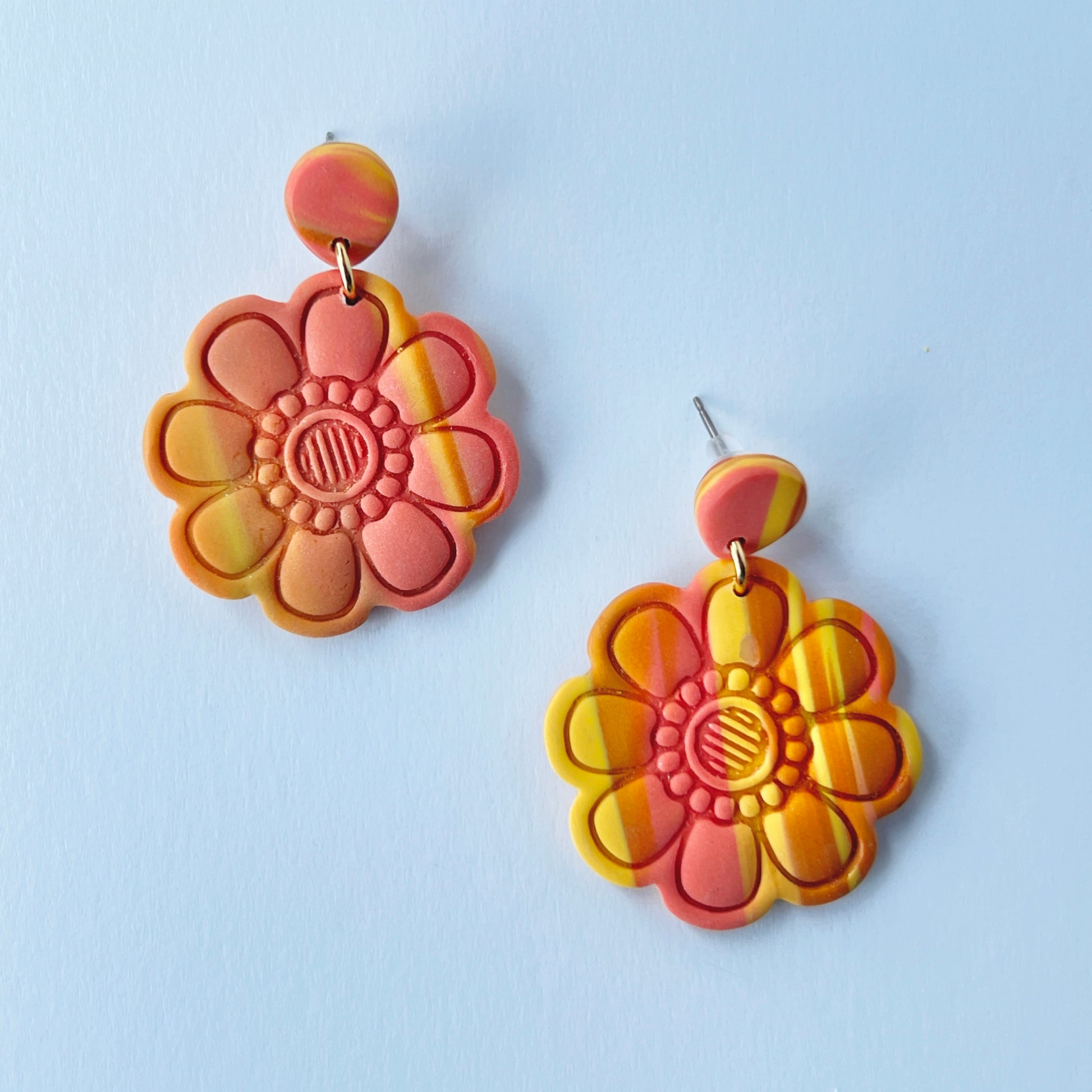 Handmade polymer clay earrings shaped like large, layered flowers with nine sculpted petals in a marbled blend of coral, orange, and yellow tones, finished with textured flower centers and coral clay stud tops on titanium posts