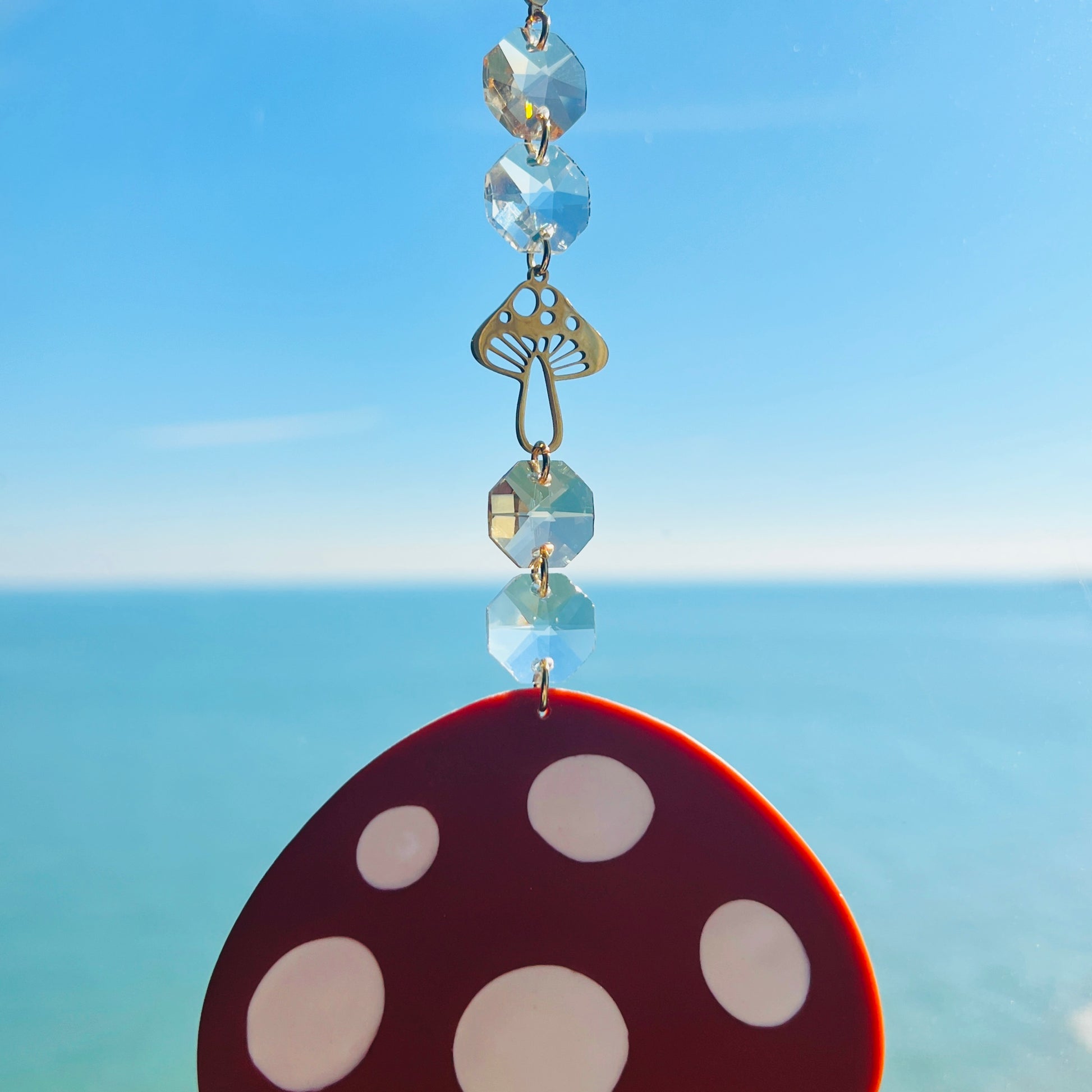 A whimsical handmade mushroom suncatcher crafted from polymer clay, featuring a brown and white spotted toadstool design suspended from a gold chain, with a dangling faceted glass crystal that casts rainbows in the sunlight. Ideal for woodland, fairycore, or cottagecore decor.