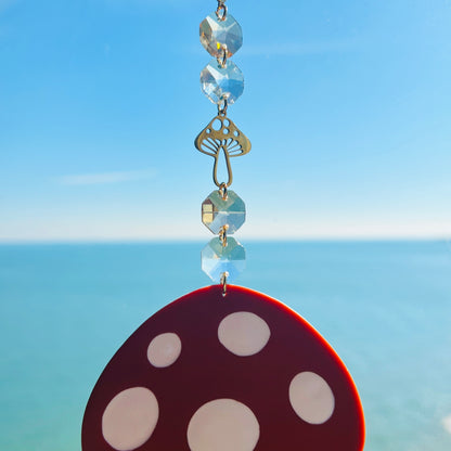 A whimsical handmade mushroom suncatcher crafted from polymer clay, featuring a brown and white spotted toadstool design suspended from a gold chain, with a dangling faceted glass crystal that casts rainbows in the sunlight. Ideal for woodland, fairycore, or cottagecore decor.