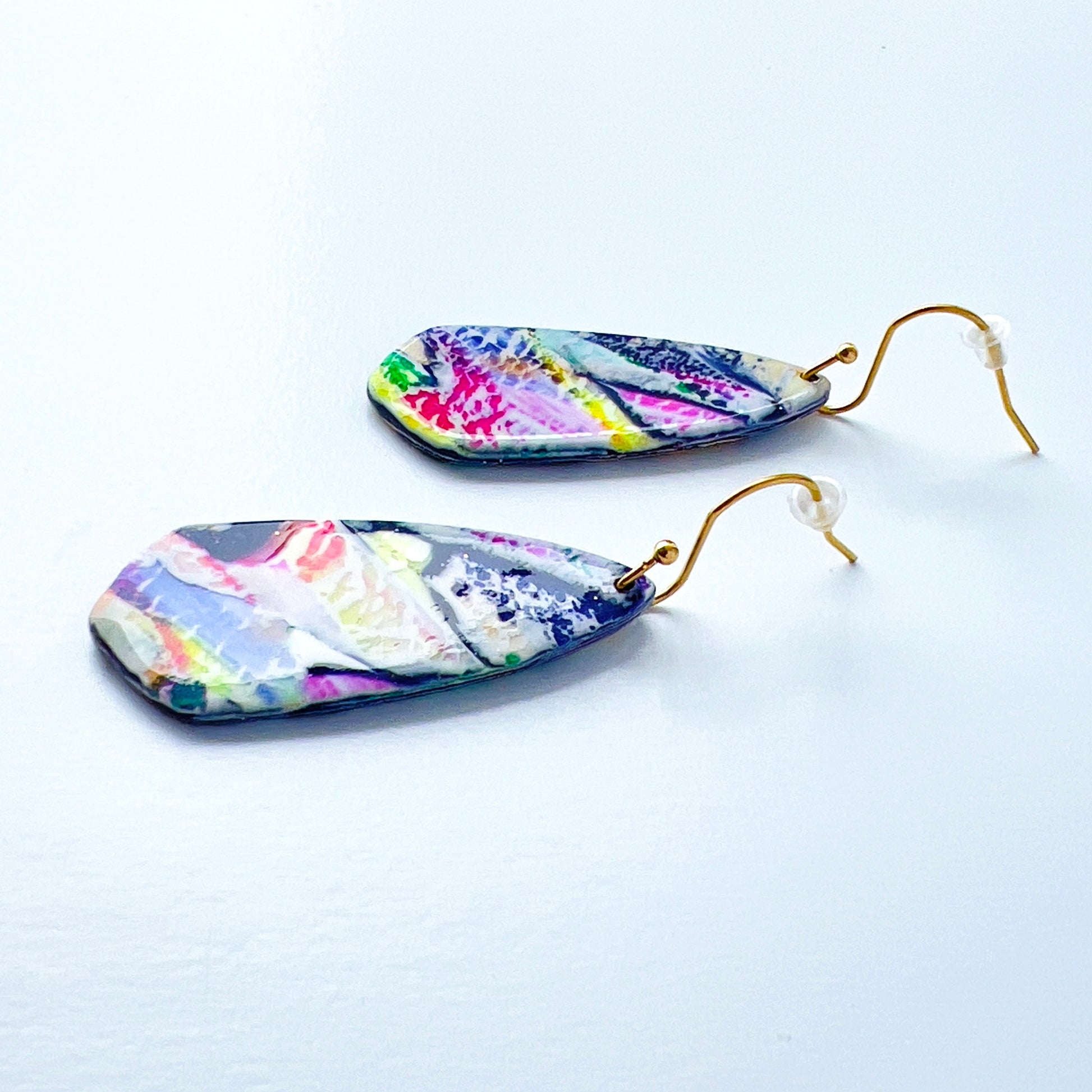 Asymmetrical dagger-shaped earrings made of polymer clay with vibrant brushstroke patterns in rainbow colors. Hanging on gold stainless steel earwires.