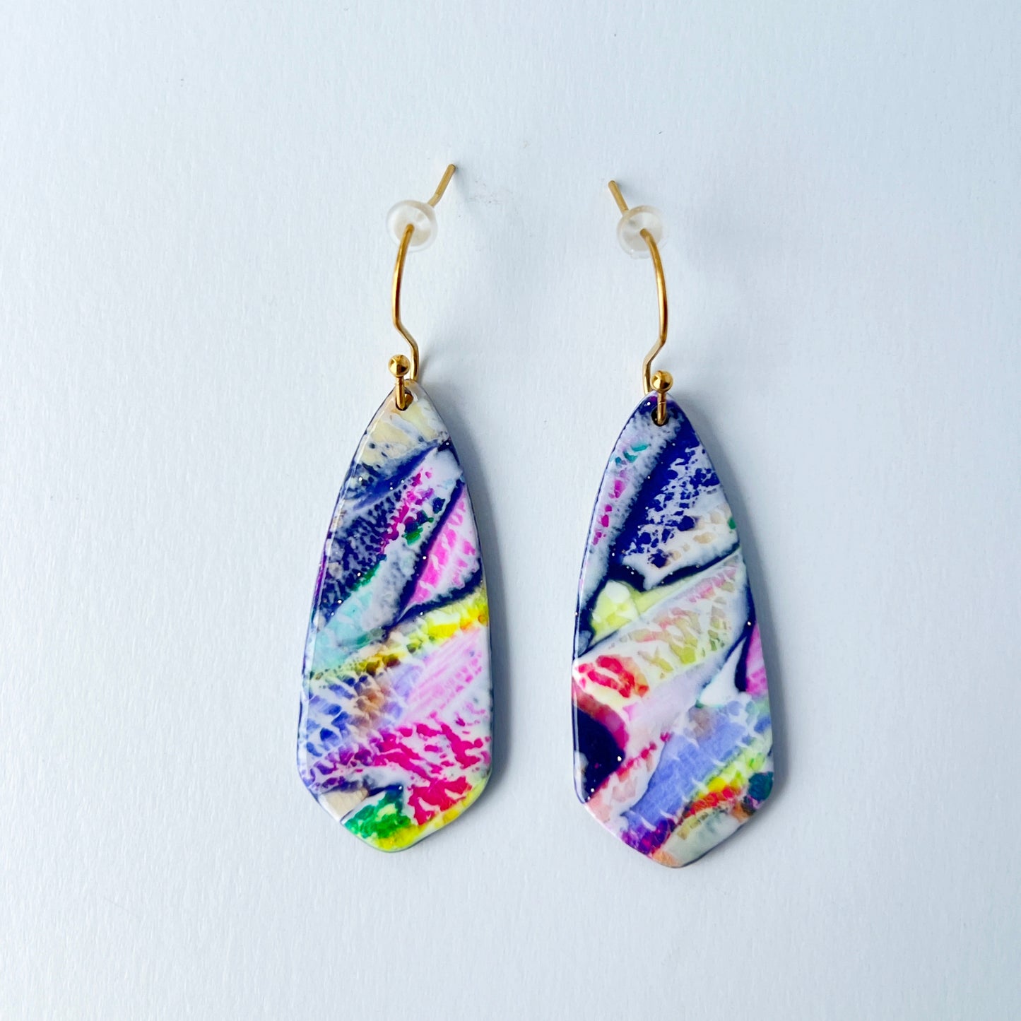 Asymmetrical dagger-shaped earrings made of polymer clay with vibrant marbling that looks like abrushstroke patterns in rainbow colors. Hanging on gold stainless steel earwires.