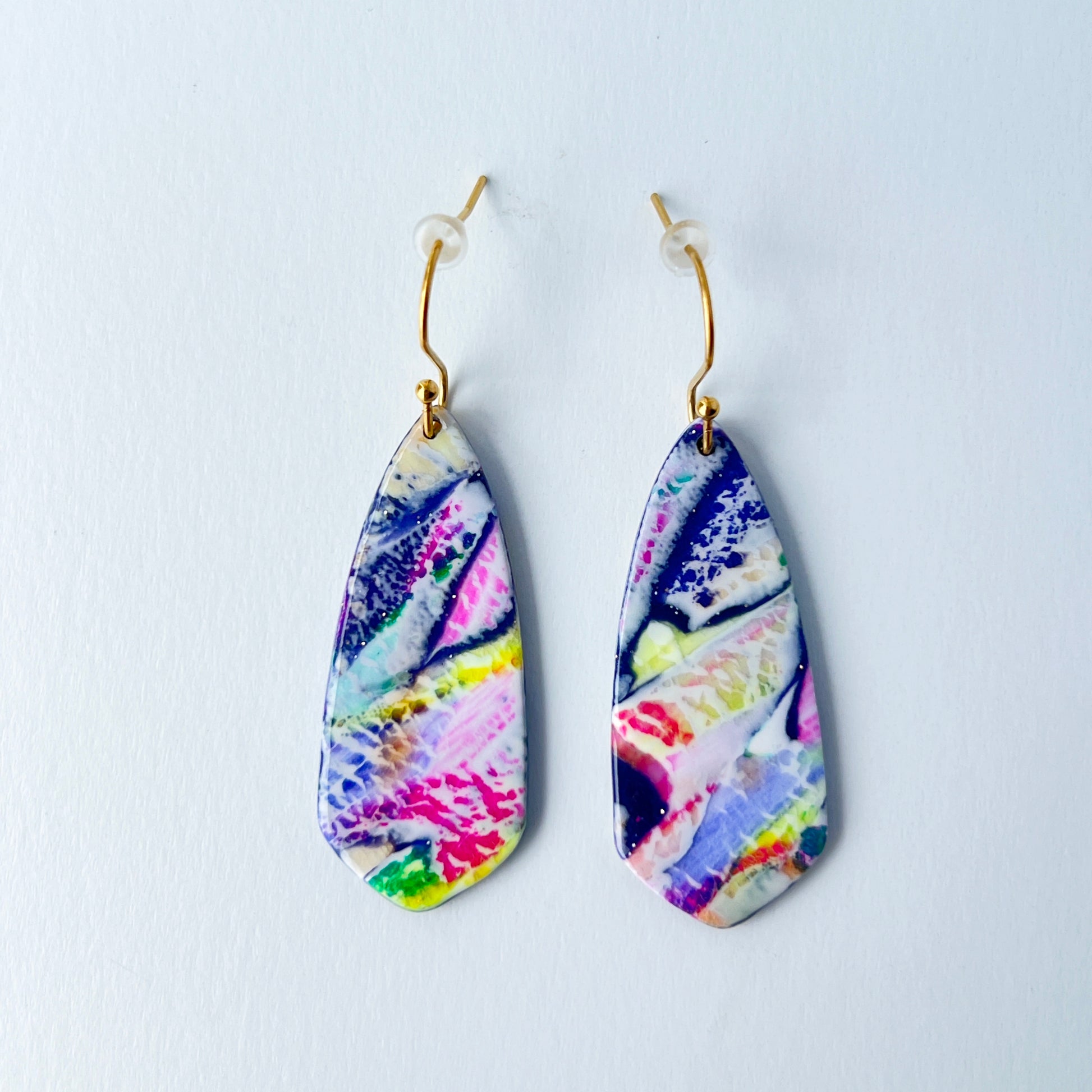 Asymmetrical dagger-shaped earrings made of polymer clay with vibrant marbling that looks like abrushstroke patterns in rainbow colors. Hanging on gold stainless steel earwires.