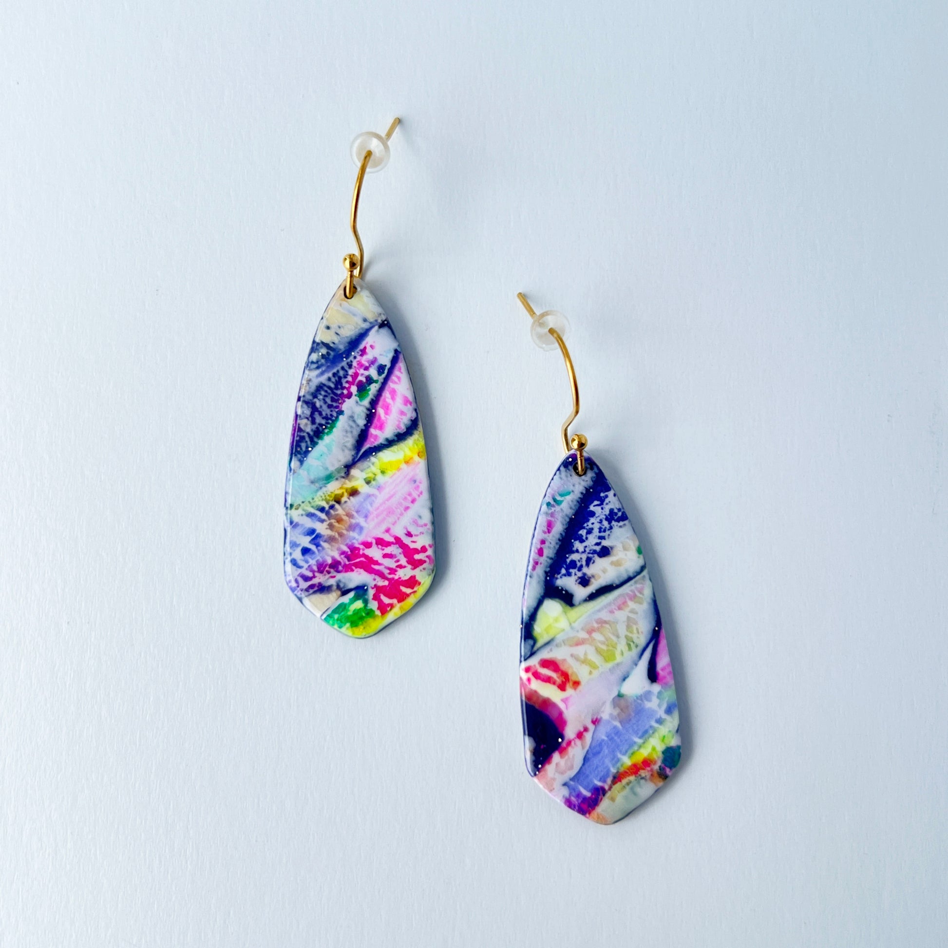 Asymmetrical dagger-shaped earrings made of polymer clay with vibrant marbling that looks like abrushstroke patterns in rainbow colors. Hanging on gold stainless steel earwires.