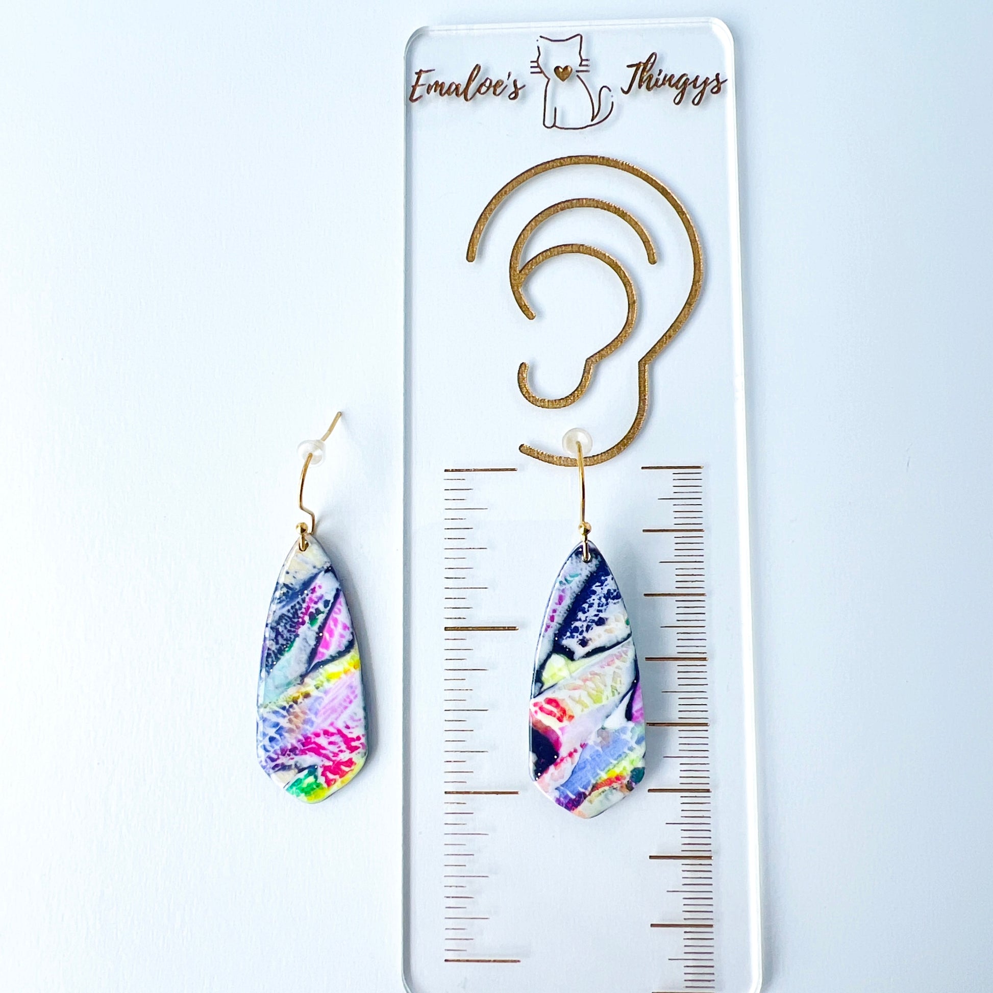 Asymmetrical dagger-shaped earrings made of polymer clay with vibrant marbling that looks like abrushstroke patterns in rainbow colors. Hanging on gold stainless steel earwires. One earring i laying on top of a clear branded ruler showing the length is 2.25 inches