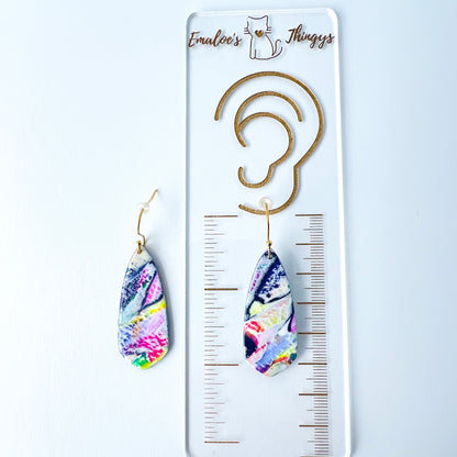 Asymmetrical dagger-shaped earrings made of polymer clay with vibrant marbling that looks like abrushstroke patterns in rainbow colors. Hanging on gold stainless steel earwires. One earring i laying on top of a clear branded ruler showing the length is 2.25 inches