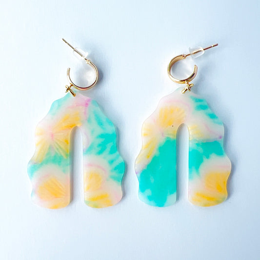 A pair of handmade polymer clay earrings shaped like arches, with swirled pastel colors and subtle details of yellow butterflies and green leafy accents. The semi-transparent clay design is paired with shiny gold-plated brass half hoop posts.