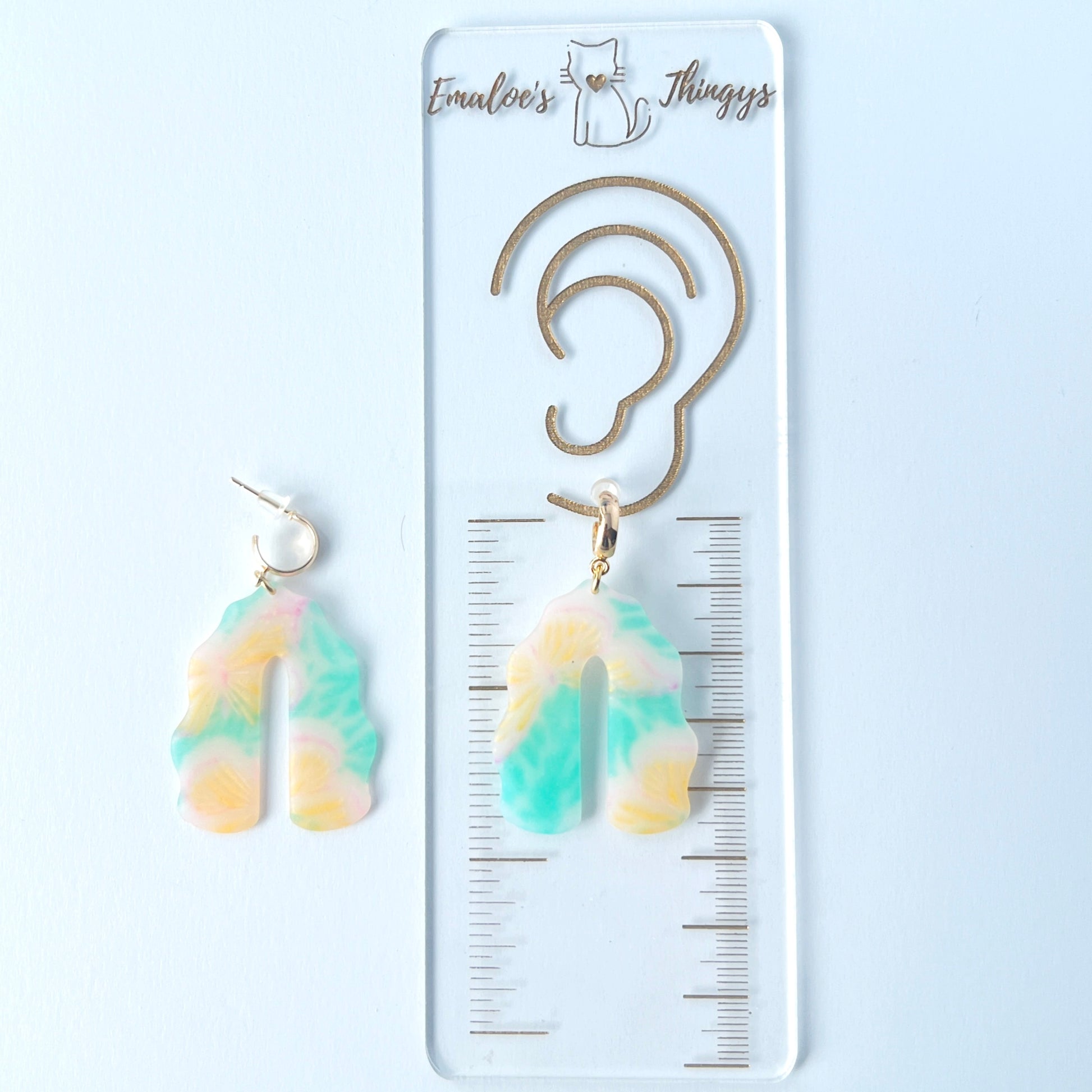 A pair of handmade polymer clay earrings shaped like arches, with swirled pastel colors and subtle details of yellow butterflies and green leafy accents. The semi-transparent clay design is paired with shiny gold-plated brass half hoop posts.  One earring is laying on a clear branded ruler showing an approximate length of 2.25 inches
