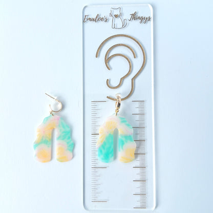 A pair of handmade polymer clay earrings shaped like arches, with swirled pastel colors and subtle details of yellow butterflies and green leafy accents. The semi-transparent clay design is paired with shiny gold-plated brass half hoop posts.  One earring is laying on a clear branded ruler showing an approximate length of 2.25 inches