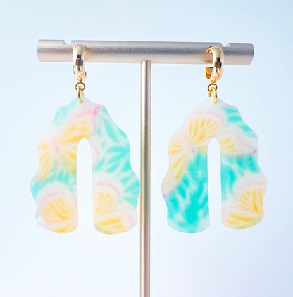 A pair of handmade polymer clay earrings shaped like arches, with swirled pastel colors and subtle details of yellow butterflies and green leafy accents. The semi-transparent clay design is paired with shiny gold-plated brass half hoop posts.
