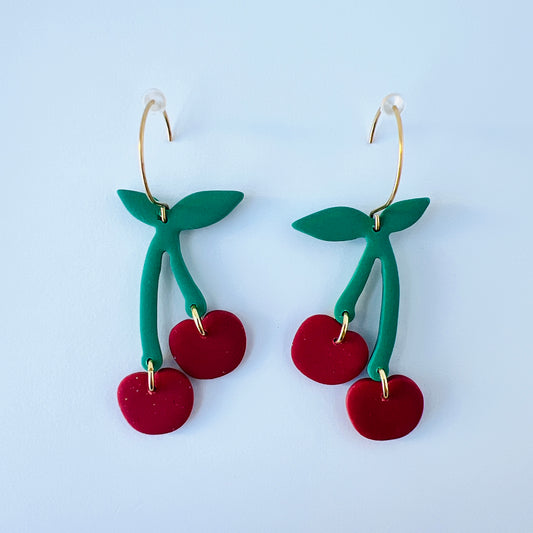 A pair of handmade cherry earrings crafted from polymer clay. Each earring features two bright red cherry shapes dangling from green leafy stems, attached to gold-plated round stainless steel earwires. The earrings are lightweight and whimsical, perfect for a fun, summery accessory.