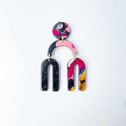 a handmade polymer clay dangle earring shaped like arched rainbows with U-shaped dangles. Marbled in a mix of pink, teal, gold, white, black, and glitter, with titanium posts. Displayed against a white background