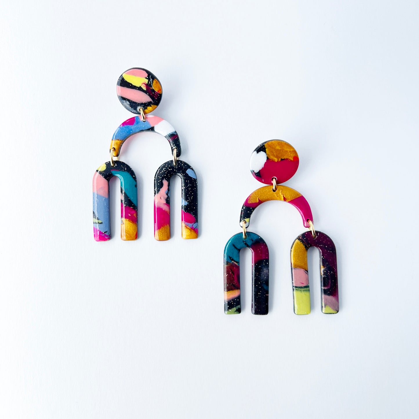 A vibrant pair of handmade polymer clay dangle earrings shaped like arched rainbows with U-shaped dangles. Marbled in a mix of pink, teal, gold, white, black, and glitter, with titanium posts. Displayed against a white background