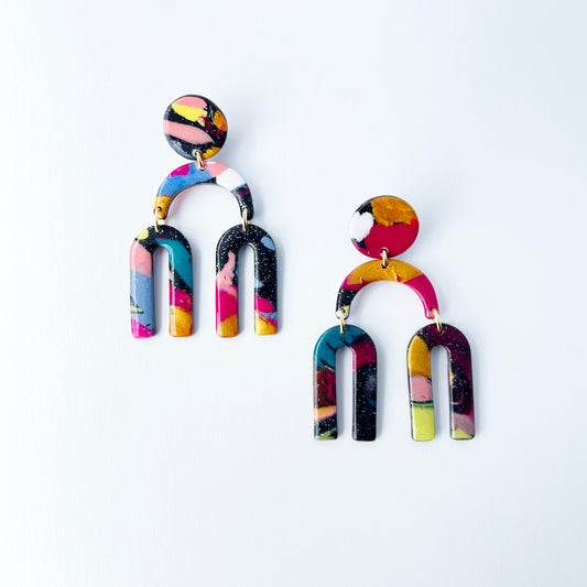 A vibrant pair of handmade polymer clay dangle earrings shaped like arched rainbows with U-shaped dangles. Marbled in a mix of pink, teal, gold, white, black, and glitter, with titanium posts. Displayed against a white background