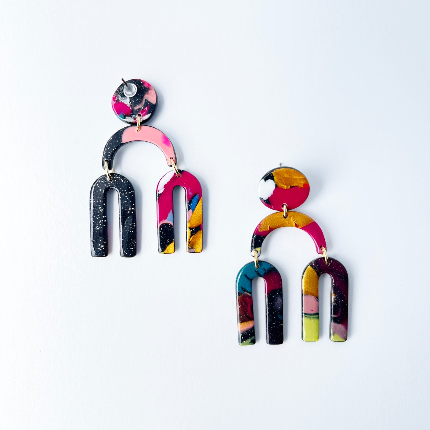 A vibrant pair of handmade polymer clay dangle earrings shaped like arched rainbows with U-shaped dangles. Marbled in a mix of pink, teal, gold, white, black, and glitter, with titanium posts. Displayed against a white background