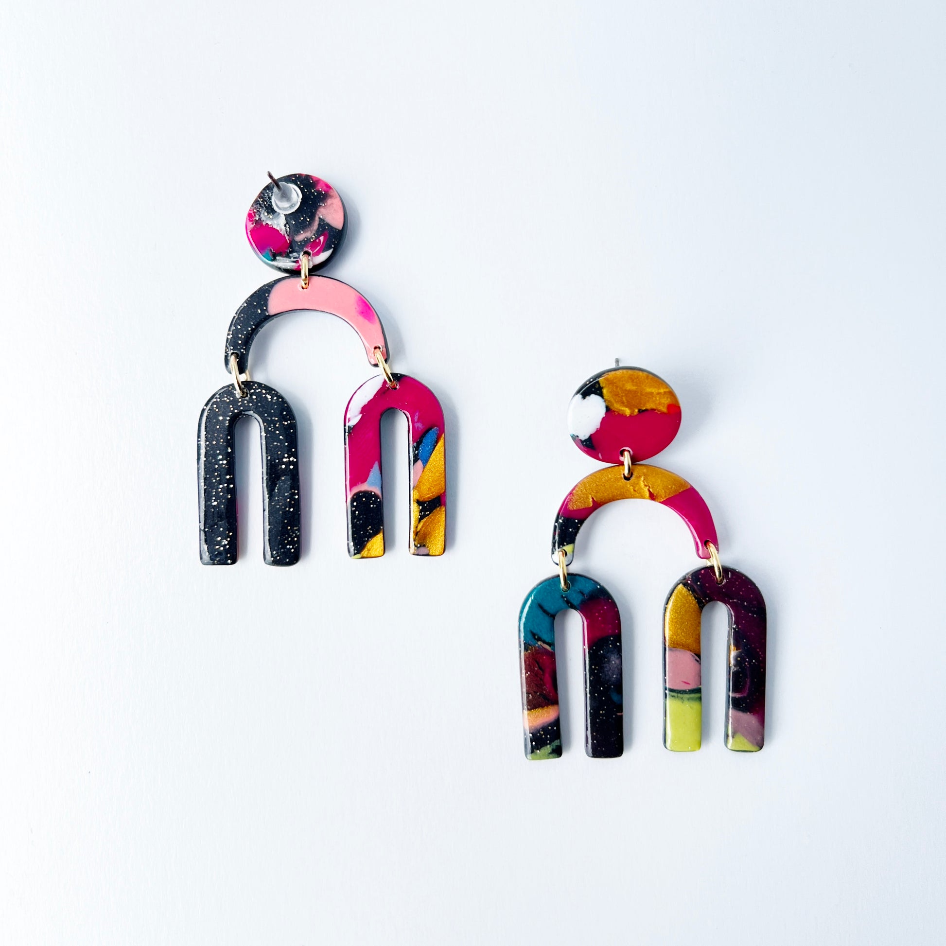 A vibrant pair of handmade polymer clay dangle earrings shaped like arched rainbows with U-shaped dangles. Marbled in a mix of pink, teal, gold, white, black, and glitter, with titanium posts. Displayed against a white background