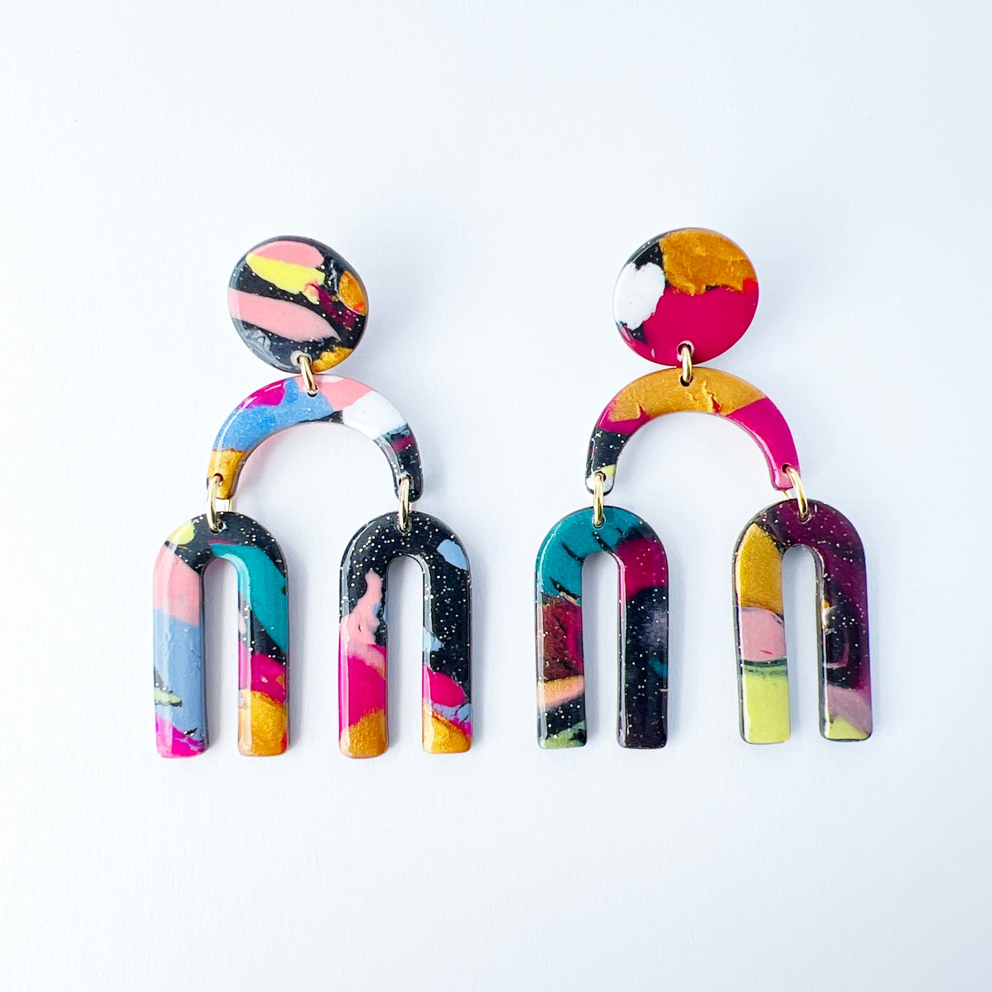 A vibrant pair of handmade polymer clay dangle earrings shaped like arched rainbows with U-shaped dangles. Marbled in a mix of pink, teal, gold, white, black, and glitter, with titanium posts. Displayed against a white background
