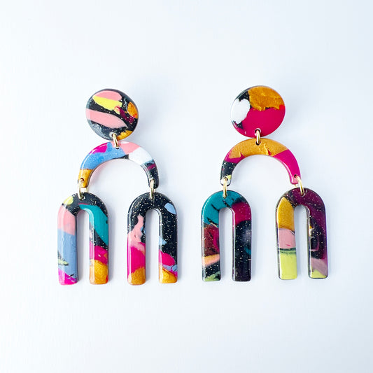 A vibrant pair of handmade polymer clay dangle earrings shaped like arched rainbows with U-shaped dangles. Marbled in a mix of pink, teal, gold, white, black, and glitter, with titanium posts. Displayed against a white background