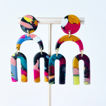 A vibrant pair of handmade polymer clay dangle earrings shaped like arched rainbows with U-shaped dangles. Marbled in a mix of pink, teal, gold, white, black, and glitter, with titanium posts. Displayed against a white background