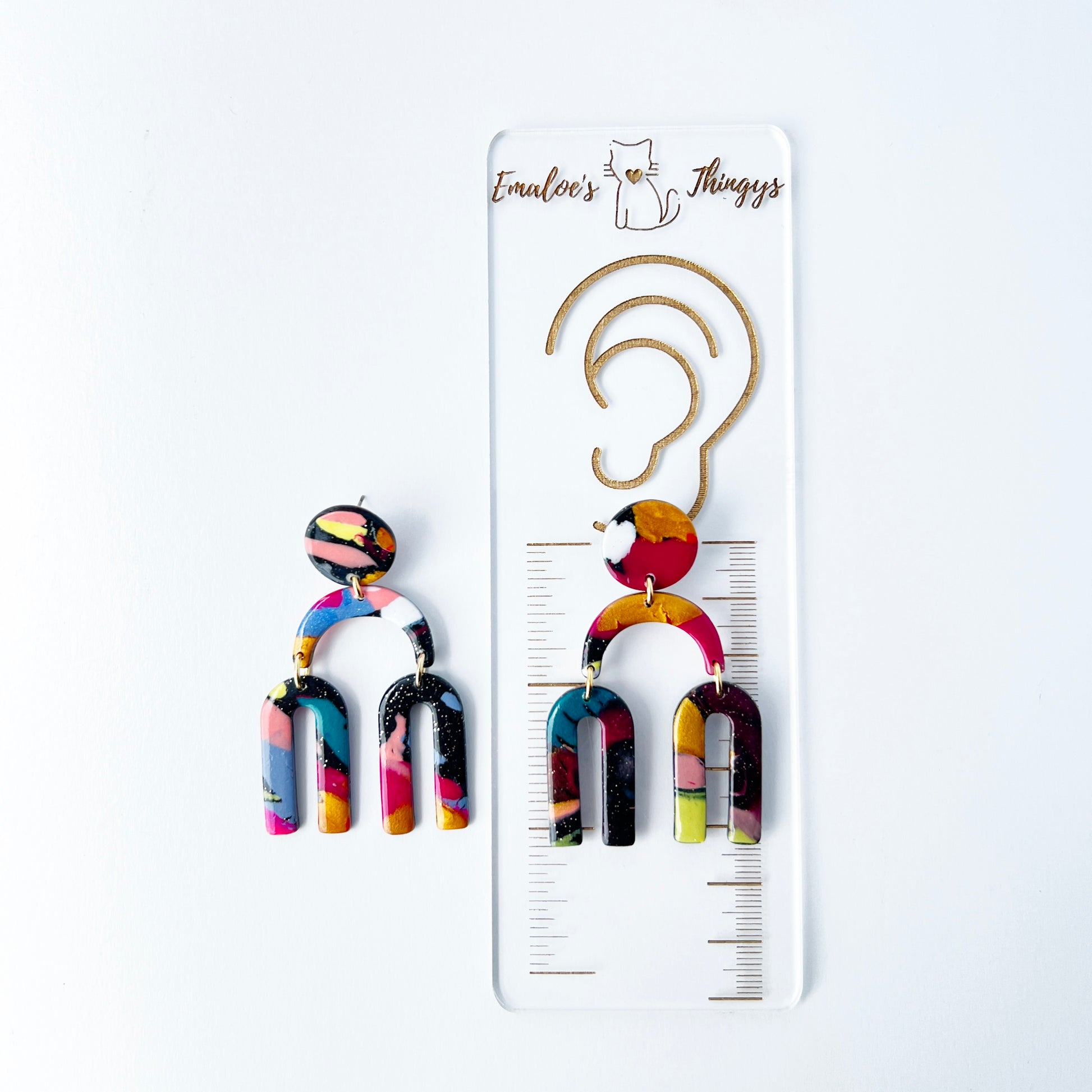 A vibrant pair of handmade polymer clay dangle earrings shaped like arched rainbows with U-shaped dangles. Marbled in a mix of pink, teal, gold, white, black, and glitter, with titanium posts. Displayed against a white background with a branded sizing ruler showing the length of the earrings are approximately 2.5 inches or 63.5mm