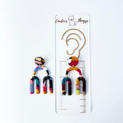 A vibrant pair of handmade polymer clay dangle earrings shaped like arched rainbows with U-shaped dangles. Marbled in a mix of pink, teal, gold, white, black, and glitter, with titanium posts. Displayed against a white background with a branded sizing ruler showing the length of the earrings are approximately 2.5 inches or 63.5mm