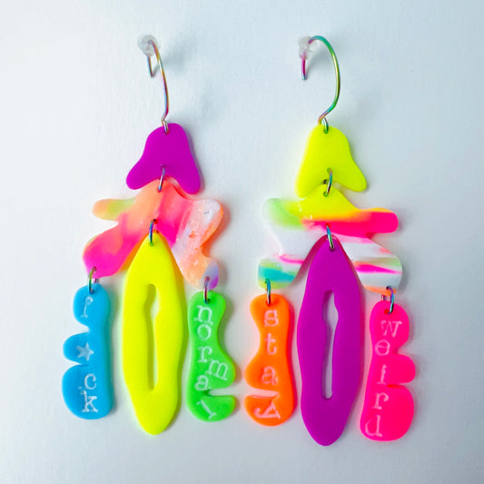 A pair of mismatched handmade statement earrings made from brightly colored neon polymer clay in layered abstract shapes. The left earring includes shapes in purple, orange, yellow, blue, and green and spells “F*ck Normal.” The right earring features neon pink, yellow, orange, purple, and green elements spelling “Stay Weird.” Both earrings are suspended from rainbow-plated stainless steel earwires and shown on a flat white background