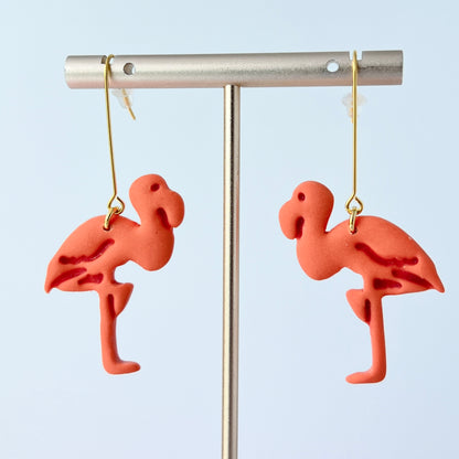 A pair of handmade coral-pink flamingo earrings sculpted from polymer clay, each suspended from long, minimalist gold-plated stainless steel bar earwires. The flamingos face each other in a mirrored design
