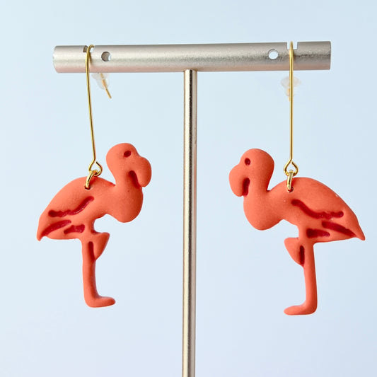 A pair of handmade coral-pink flamingo earrings sculpted from polymer clay, each suspended from long, minimalist gold-plated stainless steel bar earwires. The flamingos face each other in a mirrored design