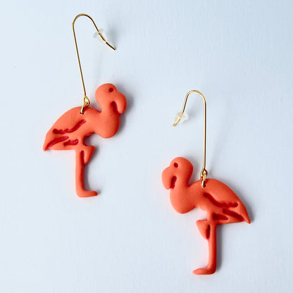 A pair of handmade coral-pink flamingo earrings sculpted from polymer clay, each suspended from long, minimalist gold-plated stainless steel bar earwires. The flamingos face each other in a mirrored design