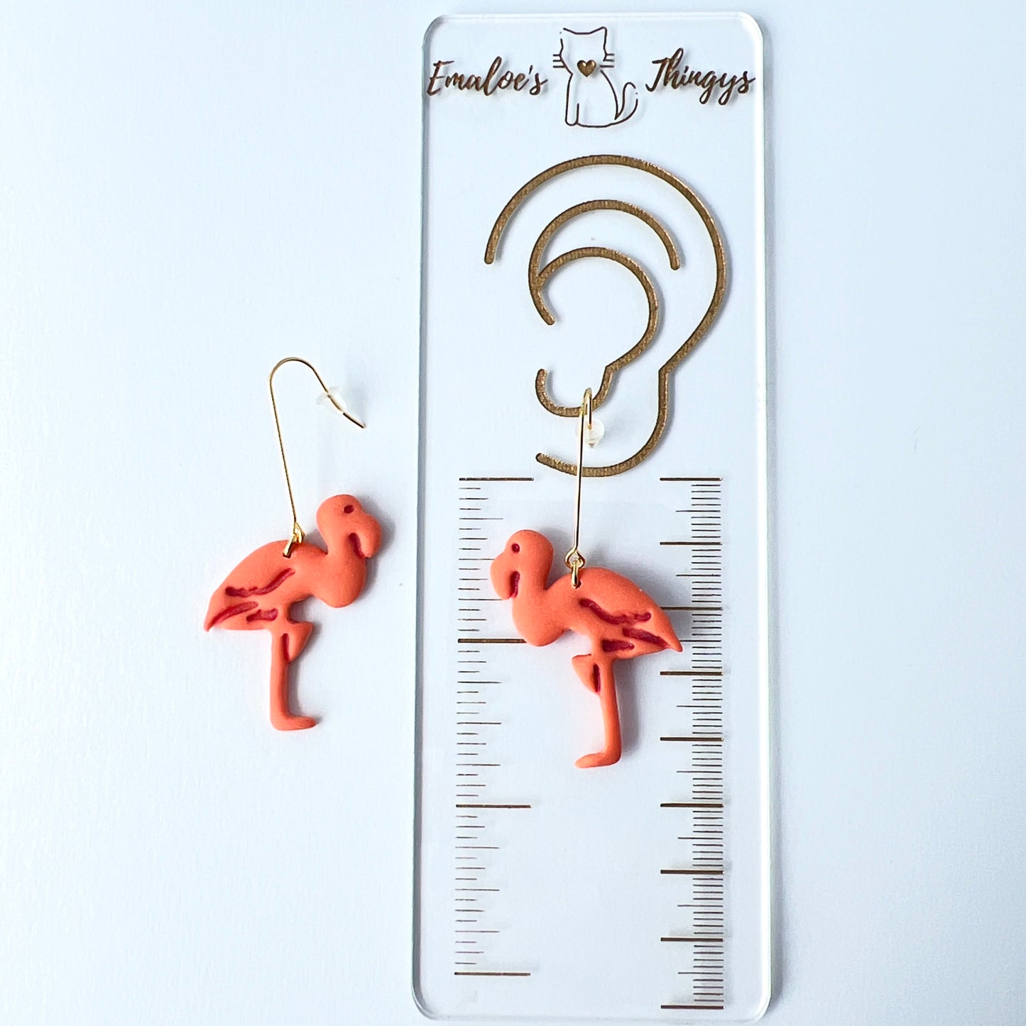 A pair of handmade coral-pink flamingo earrings sculpted from polymer clay, each suspended from long, minimalist gold-plated stainless steel bar earwires. The flamingos face each other in a mirrored design. one earring is on a card with a ruler for scale showing length of 2.5 inches