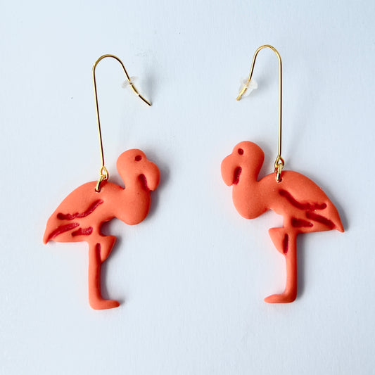 A pair of handmade coral-pink flamingo earrings sculpted from polymer clay, each suspended from long, minimalist gold-plated stainless steel bar earwires. The flamingos face each other in a mirrored design