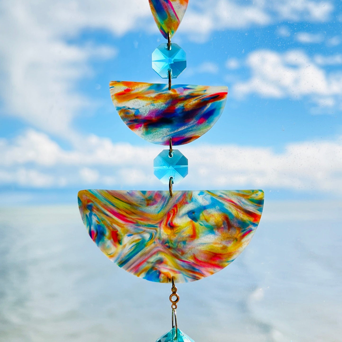 Golden Swirl: Swirled warm-toned polymer clay teardrop and arches with aqua glass crystals and a gold chain, photographed against a bright sky over water.