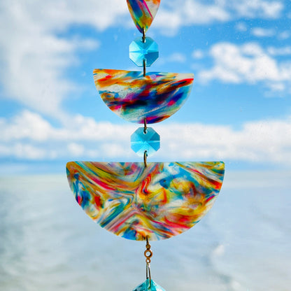 Golden Swirl: Swirled warm-toned polymer clay teardrop and arches with aqua glass crystals and a gold chain, photographed against a bright sky over water.