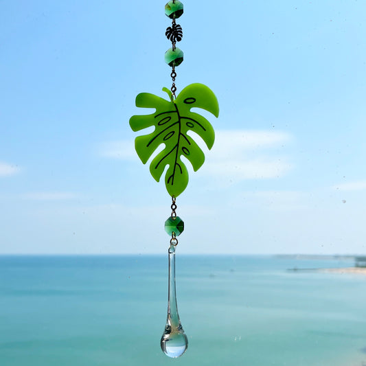 A handmade monstera leaf suncatcher made from green polymer clay with detailed vein textures, hanging from a gold loop and chain, featuring a faceted glass crystal that casts rainbows in sunlight. Ideal for botanical or boho home decor.