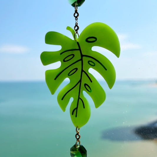 A handmade monstera leaf suncatcher made from green polymer clay with detailed vein textures, hanging from a gold loop and chain, featuring a faceted glass crystal that casts rainbows in sunlight. Ideal for botanical or boho home decor.