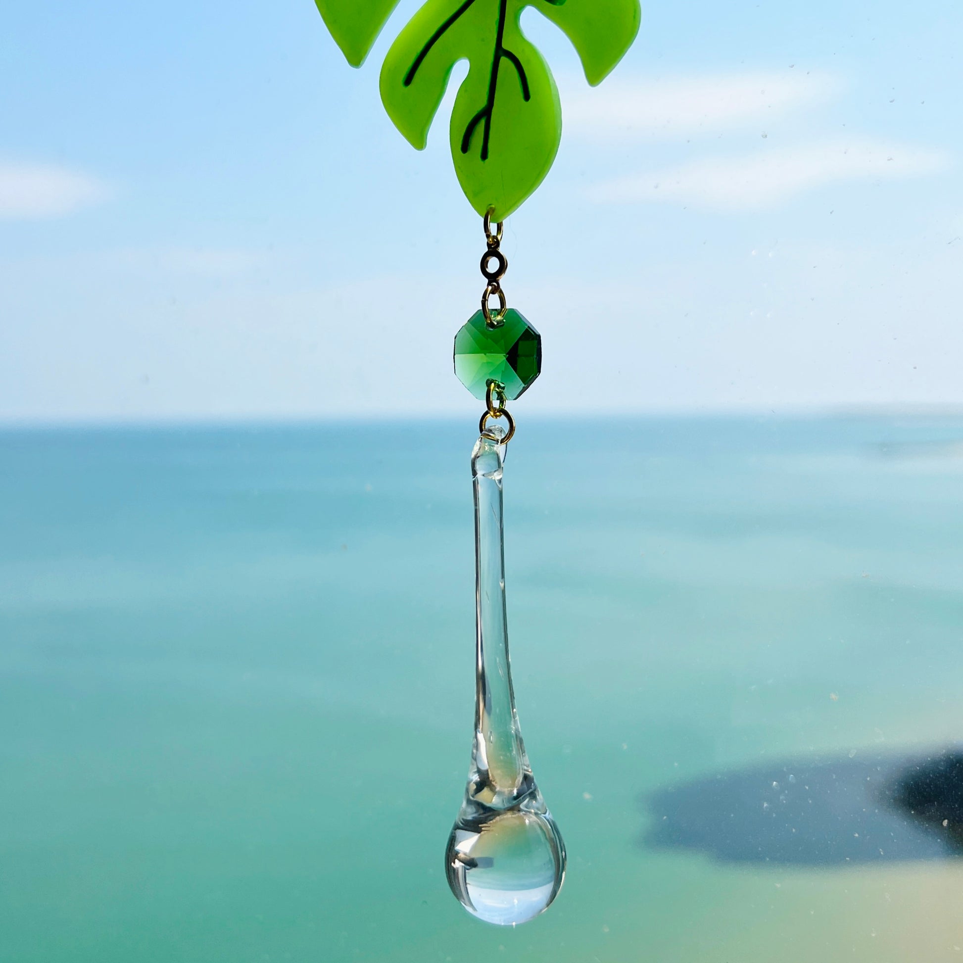 A handmade monstera leaf suncatcher made from green polymer clay with detailed vein textures, hanging from a gold loop and chain, featuring a faceted glass crystal that casts rainbows in sunlight. Ideal for botanical or boho home decor.