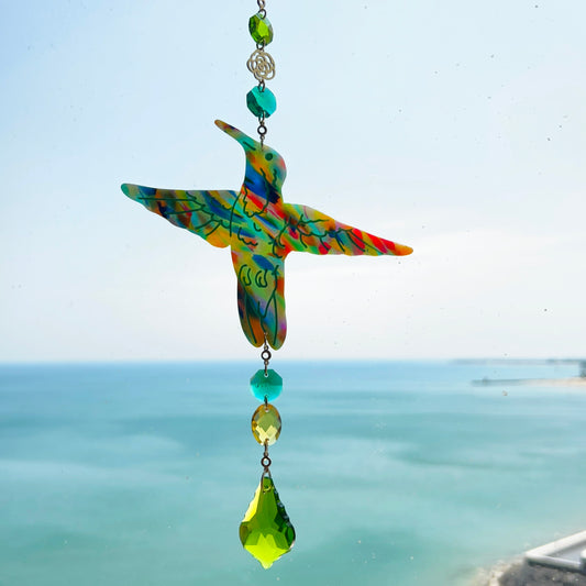 A handmade hummingbird suncatcher crafted from colorful polymer clay, featuring a detailed hummingbird design in shades of green, blue, and pink, suspended from a gold chain with a faceted green maple leaf glass crystal that casts rainbows in the sunlight. Perfect for nature lovers and whimsical window decor.