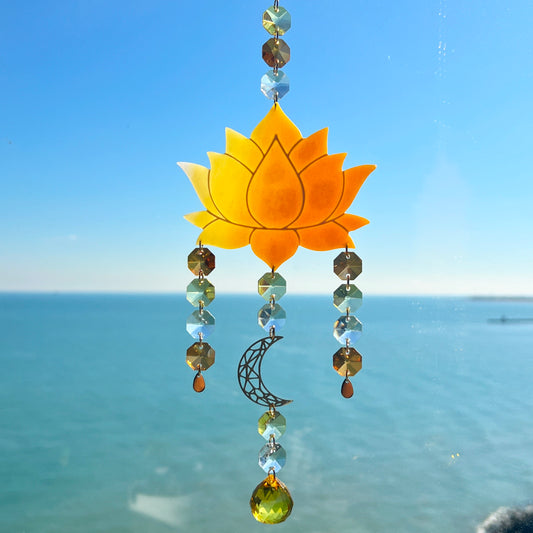 A handmade lotus flower suncatcher crafted from ombre orange & yellow polymer clay, featuring a stylized lotus bloom suspended from a gold chain with a faceted glass crystal hanging below, designed to reflect rainbow light in the sun. Ideal for meditation rooms, spiritual decor, or peaceful window displays.