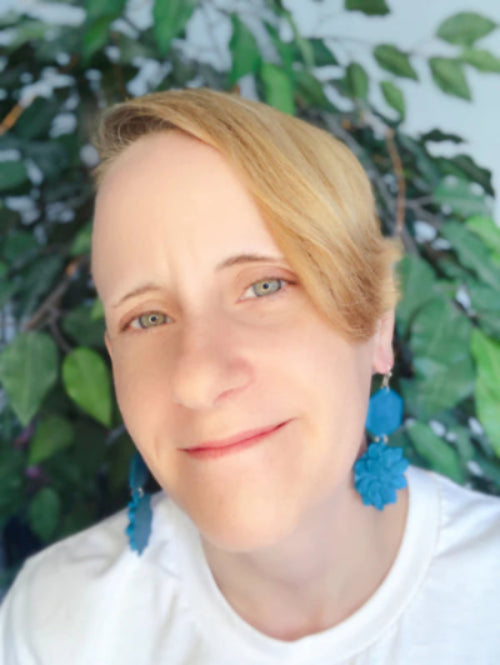 An image of the Maker - Jenn with short blonde hair wearing blue earrings against a green leafy background
